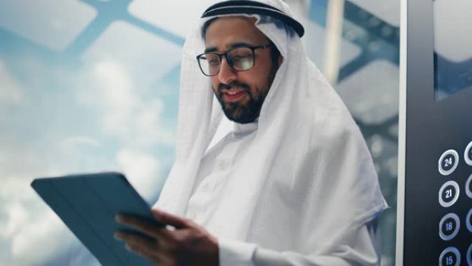 成功的穆斯林商人在传统的白色坎杜拉骑着玻璃电梯到现代商务中心的办公室。使用平板电脑的人。沙特、阿联酋