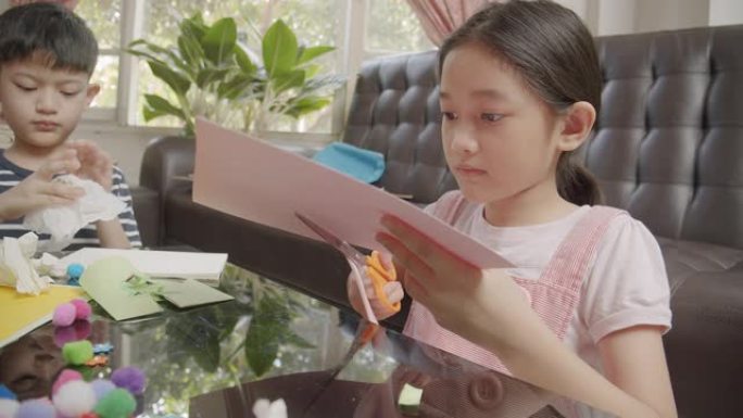 两个亚洲同胞男孩和女孩在家里的同时在客厅剪纸并制作手工艺品。他们使用彩纸，剪刀和胶水在纸上创作艺术作