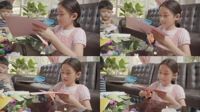 两个亚洲同胞男孩和女孩在家里的同时在客厅剪纸并制作手工艺品。他们使用彩纸，剪刀和胶水在纸上创作艺术作