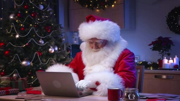 体贴的圣诞老人坐在他的笔记本电脑上，坐在装饰精美的厨房的桌子上，身后有一棵圣诞树