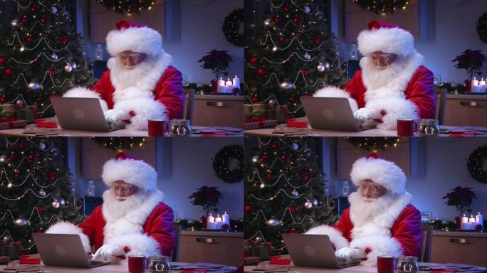 体贴的圣诞老人坐在他的笔记本电脑上，坐在装饰精美的厨房的桌子上，身后有一棵圣诞树