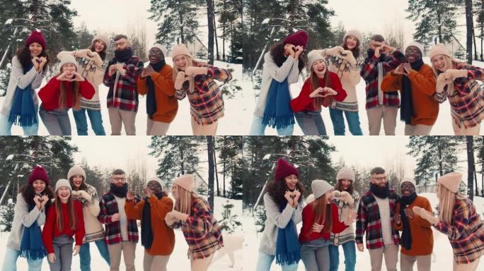 团结概念。快乐的多民族朋友看着相机，在白雪皑皑的森林慢动作中展示爱心手势。