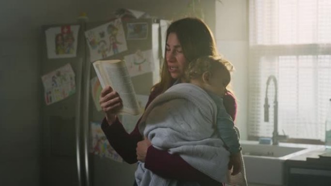 年轻的快乐和平母亲的电影镜头正在读童话故事给蹒跚学步的男婴，同时在家里的厨房里睡在怀里。美梦的概念，