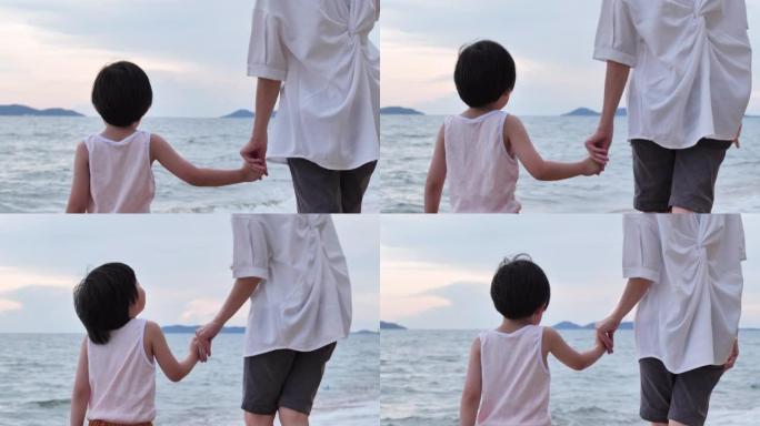 亚洲4岁母亲和儿子的家庭的背景图，他带着母亲散步，在沙滩上沿着日落海浪的边缘度过时光。度假概念。
