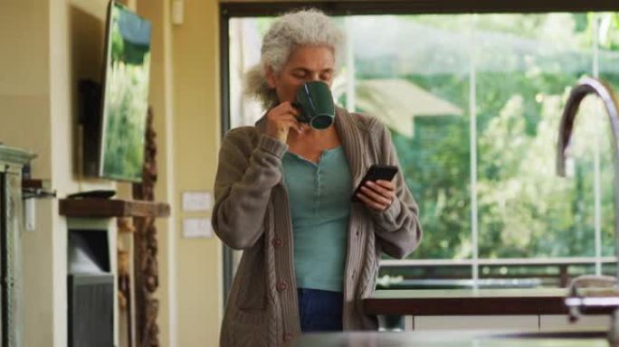 高级混血妇女在厨房喝咖啡和使用智能手机的肖像