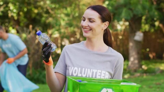 微笑的白人妇女穿着志愿者t恤，拿着回收板条箱，收集塑料垃圾