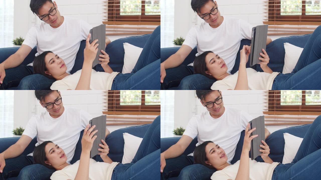 亚洲夫妇在家中客厅使用平板电脑在互联网上进行在线购物，甜蜜的夫妇在家中放松时躺在沙发上享受爱情时刻。