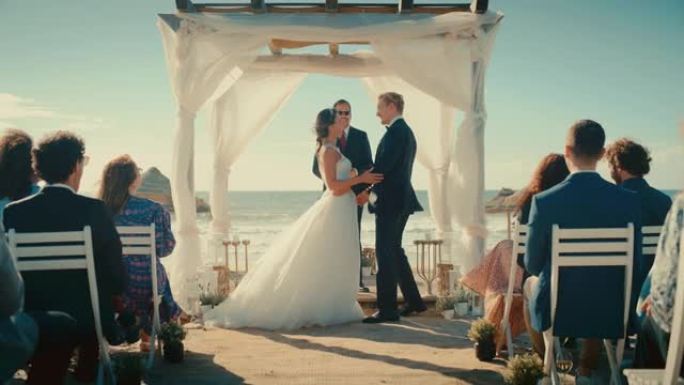 美丽的新娘和新郎在海边的海滩上举行户外婚礼。浪漫情侣结婚、接吻和拥有多民族文化的朋友庆祝的完美场所。