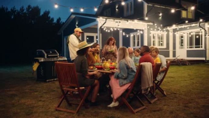 父母，孩子和朋友聚集在一个带有灯光装饰的美丽家外的烧烤餐桌旁。老人和年轻人玩得开心，吃饭。后院的花园
