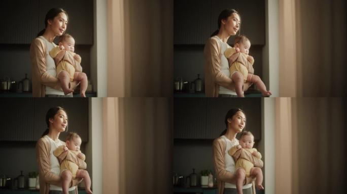 美丽的年轻亚洲母亲抱着婴儿，站在家里的窗户旁边。快乐的女人，刚接触母亲，微笑着，和蹒跚学步的孩子一起