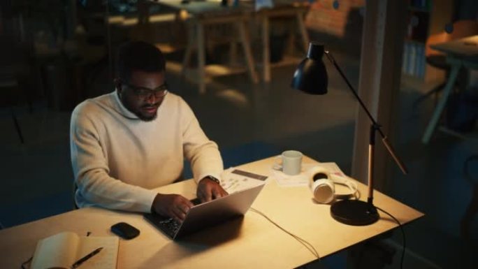 时尚的黑人专家晚上坐在创意机构的办公桌前。年轻英俊的非洲男子在营销公司的笔记本电脑上工作。静态高角度