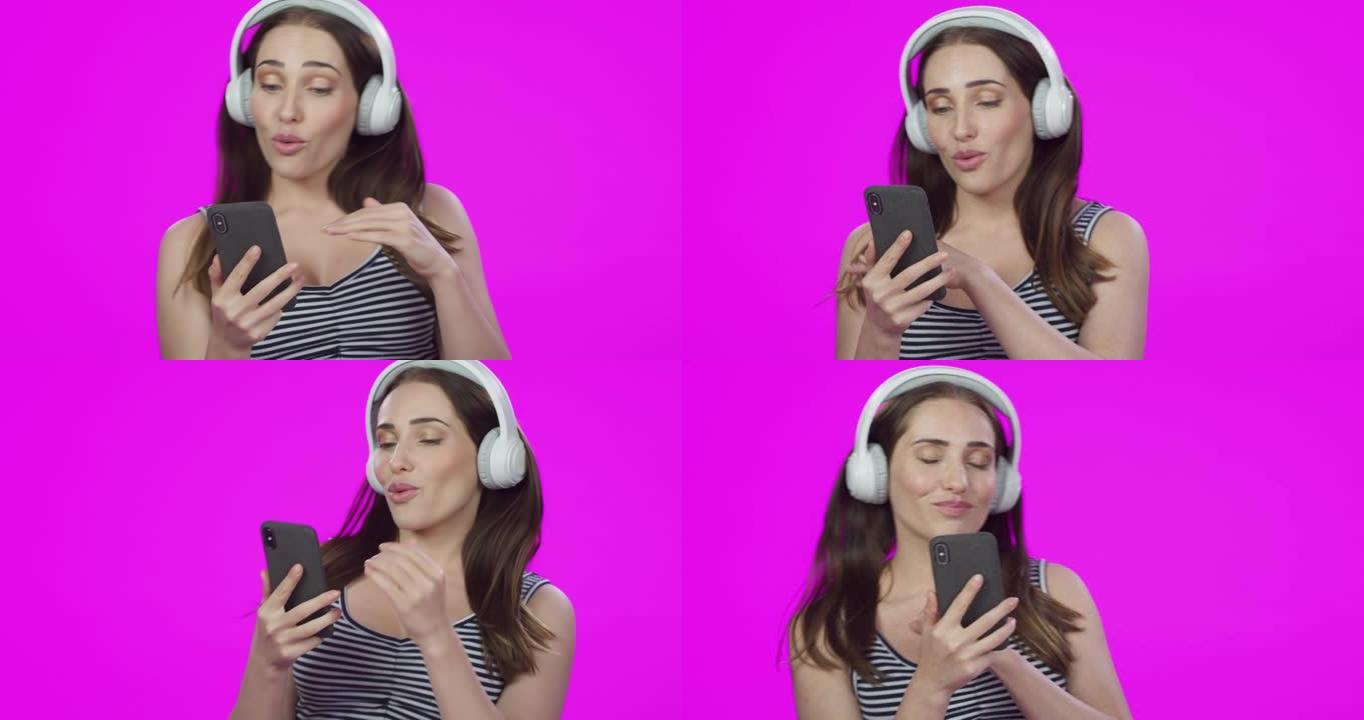 4k视频片段，一个美丽的年轻女子使用带智能手机的耳机，在粉红色的工作室背景下跳舞