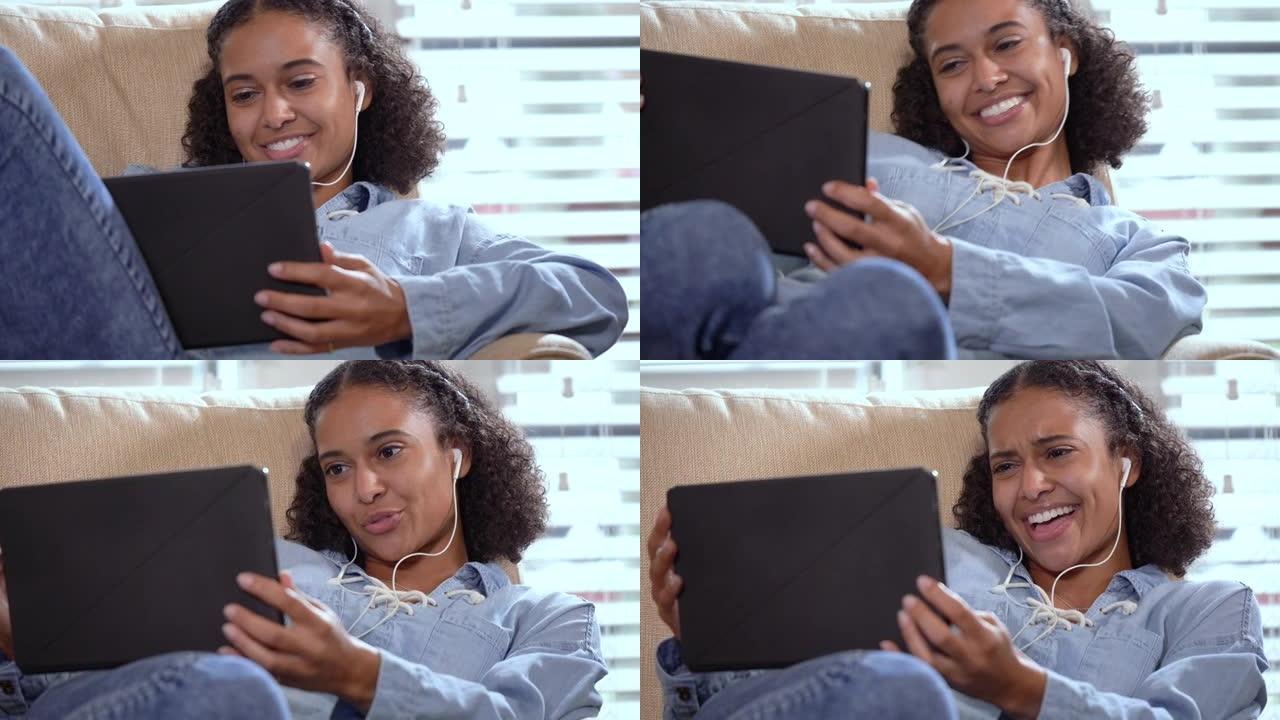 使用平板电脑，耳塞坐在扶手椅上的年轻混血妇女
