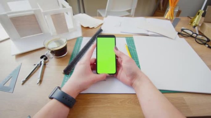 建筑师坐在办公室的办公桌上的POV镜头，使用带有绿色屏幕的手机进行计划-慢动作拍摄