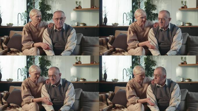 忧心忡忡的65-70岁退休的高级妻子坐在舒适的家庭客厅的沙发上与丈夫谈论他的健康。