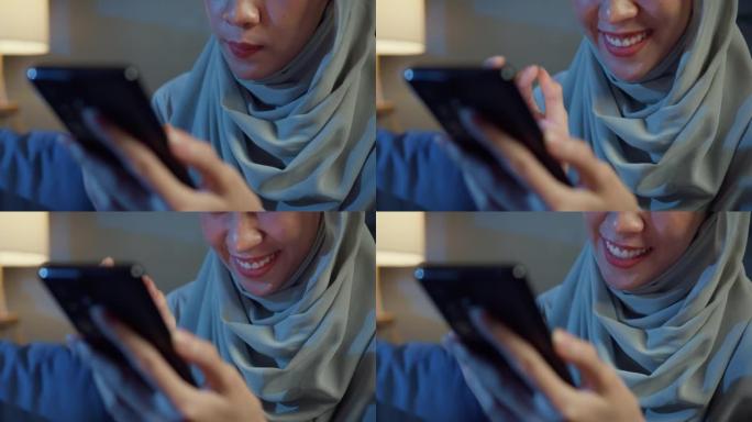 特写迷人开朗的年轻亚洲穆斯林美女戴着头巾，随意坐在沙发上，晚上在家里的客厅享受网上购物。女孩伊斯兰生
