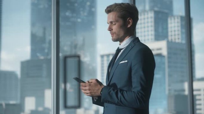 自信的年轻商人穿着西装站在现代办公室，使用智能手机，看着窗外的摩天大楼。成功的财务经理计划工作项目。