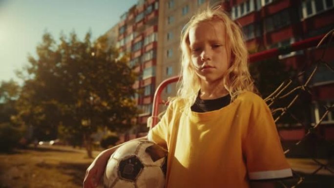 一个穿着黄色t恤的严肃的金发女孩的肖像，她在附近拿着一个足球，站在球门门旁边。年轻的足球运动员看着相