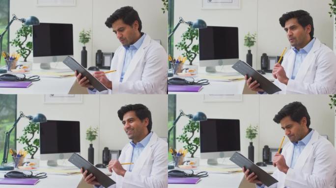 男性医生或全科医生穿着白大褂思考坐在办公室的办公桌上在剪贴板上写笔记-慢动作拍摄