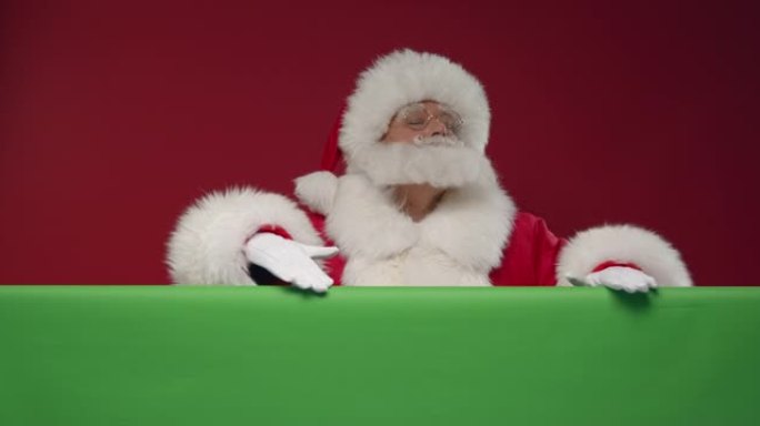 圣诞老人从他面前的绿色屏幕后面出现在红色背景的框架中，用手摩擦屏幕，竖起大拇指并走开，