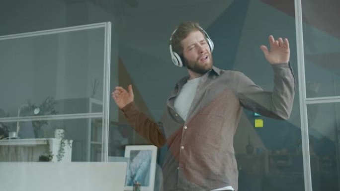 顽皮的员工有魅力的男人戴着耳机在玻璃墙办公室跳舞