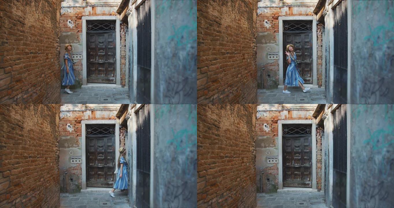 侧视图年轻快乐快乐的金发女游客走过，在威尼斯狭窄的老城区通道对着镜头挥手致意。