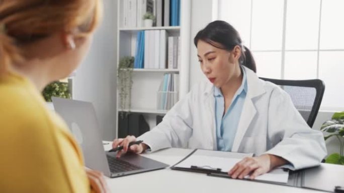 年轻的亚洲女性医生在白色医疗制服使用笔记本电脑正在传递伟大的新闻谈话讨论结果或症状与女孩患者坐在办公