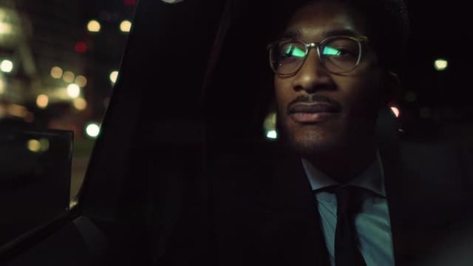 穿着眼镜的时髦黑人的肖像晚上正坐在出租车的后座上通勤回家。商业首席执行官看着窗外，在温暖的灯光下，在