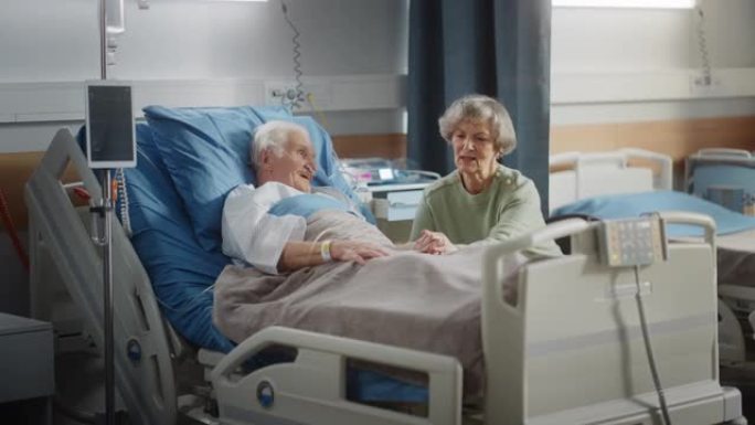 医院病房: 老人躺在床上休息，他有爱心的美丽妻子来访，支持他坐在旁边，手牵着手，幸福的夫妻面带微笑。