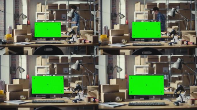 台式计算机显示器站在桌子上，带有绿屏Chromakey模拟显示器。小型企业仓库，工人在后台行走。带纸