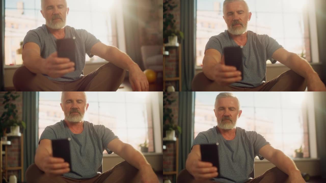 在阳光公寓的家中，在晨练期间，在锻炼之间使用智能手机的强壮健康的高级男子的特写肖像。健康的生活方式，