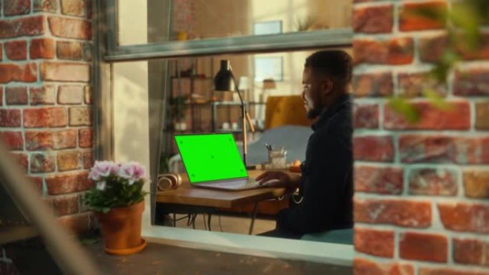 非洲男子坐在时尚客厅的桌子旁，在一台带有绿屏模拟显示的笔记本电脑上工作。业务经理在家远程工作。从窗外