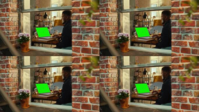 非洲男子坐在时尚客厅的桌子旁，在一台带有绿屏模拟显示的笔记本电脑上工作。业务经理在家远程工作。从窗外