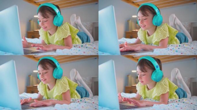 卧室里的女孩躺在床上，戴着无线耳机在笔记本电脑上听音乐或流媒体电影-慢动作拍摄