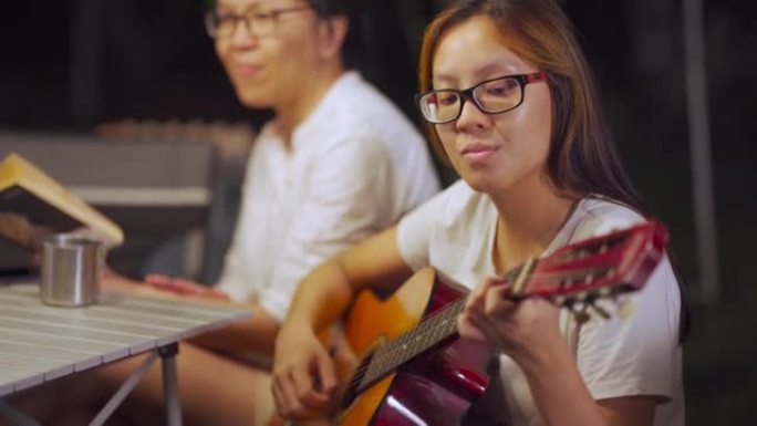 亚洲华裔女儿与家人和朋友哼着吉他，晚上在花园聚会露营帐篷中享受
