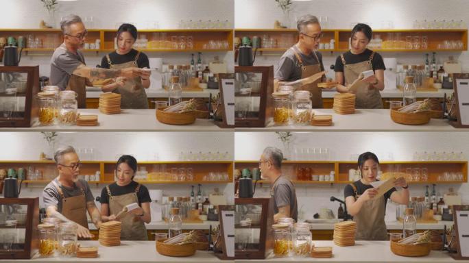 亚洲华人高级男咖啡馆老板在咖啡店柜台安排杯垫时向女儿展示食物菜单