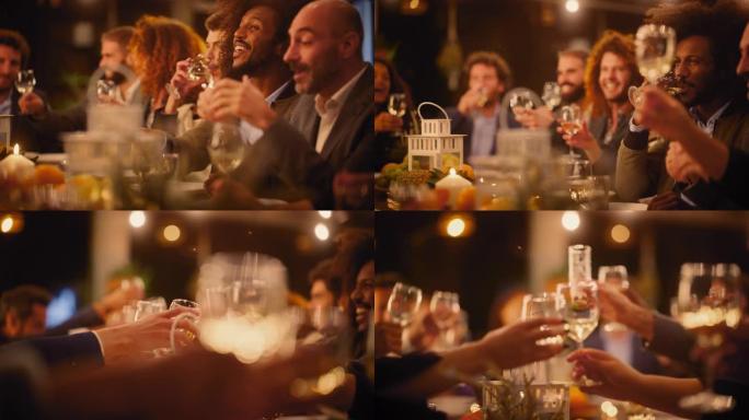 大型晚宴，一小群多种族的不同朋友在餐厅庆祝。美丽快乐的主人在晚上坐在桌子旁敬酒并举起酒杯。