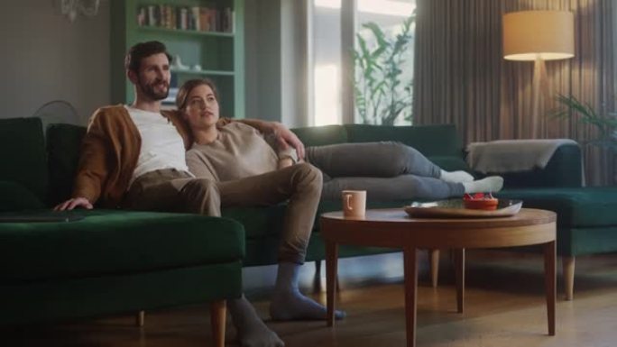 夫妇坐在客厅的沙发上看电视。女朋友和男朋友躺在一起拥抱，用遥控器切换频道。在舒适时尚的室内一起在家度