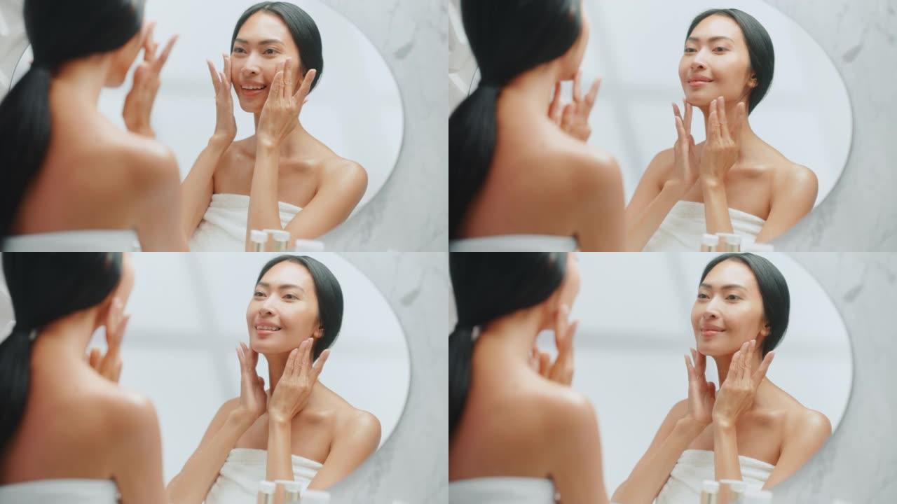 美丽的亚洲女人的肖像轻轻地涂面霜看着浴室的镜子。年轻女性用天然抗衰老化妆品护肤品让她的皮肤柔软、光滑