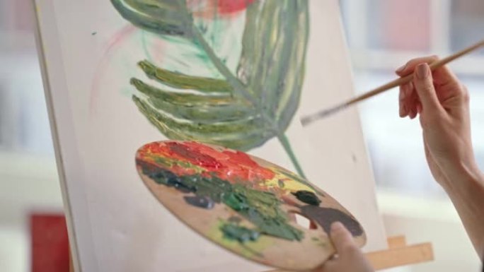 年轻的女画家在工作室里在画布上画彩画。艺术家在车间用画笔画一朵花。女人在僻静的商店里画玫瑰的图像