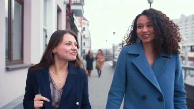 宽容和友谊。两个年轻快乐美丽的多民族女性朋友一起走在城市街道上聊天。