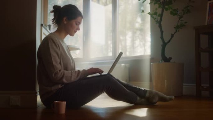 年轻女子在家使用笔记本电脑进行远程工作。美丽的女孩坐在地板上使用互联网，喝早晨的一杯咖啡。阳光明媚的