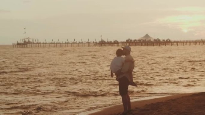 年轻的父亲抱着小儿子一起度假，在海边海浪上看着迷人的夏日日落景色。