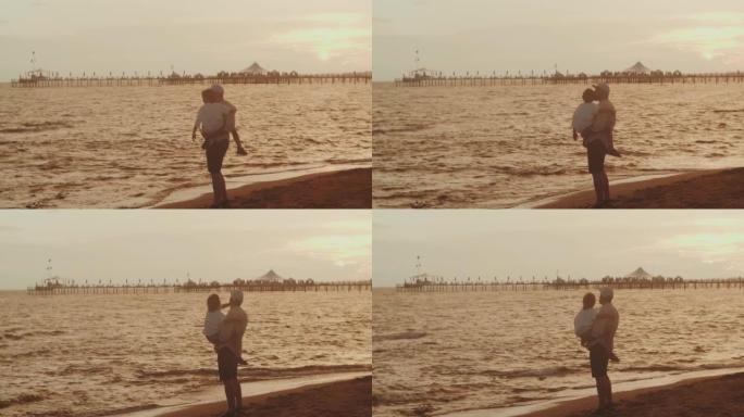 年轻的父亲抱着小儿子一起度假，在海边海浪上看着迷人的夏日日落景色。