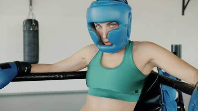 休息时戴着头盔手套和运动胸罩站在环形角的女拳击手的慢动作肖像