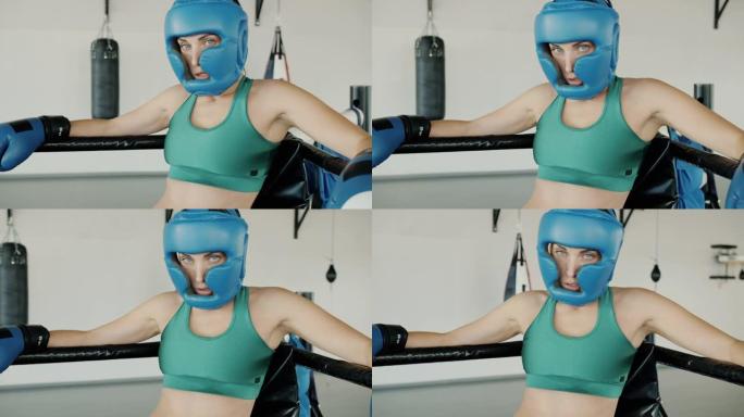 休息时戴着头盔手套和运动胸罩站在环形角的女拳击手的慢动作肖像