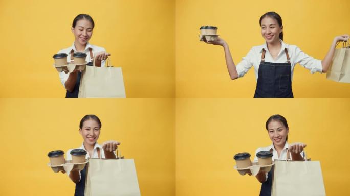 快乐微笑亚洲女服务员穿着围裙友好地看着相机，用工艺纸袋给孤立在黄色背景上的顾客咖啡或茶和美味的食物。