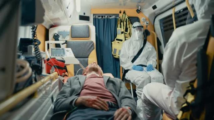 穿着一次性工作服的专业EMS护理人员团队与受伤的年轻男性患者一起乘坐救护车。急诊助理坐在去医院的路上