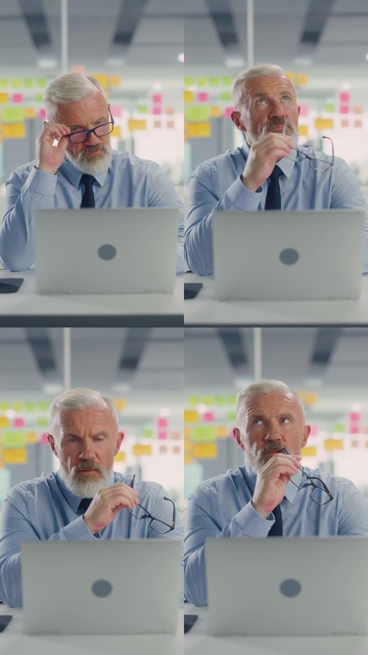 一个忙碌的高级商人在现代办公室使用笔记本电脑的垂直肖像。经理休息了眼睛，思考成功的财务想法。压力很大