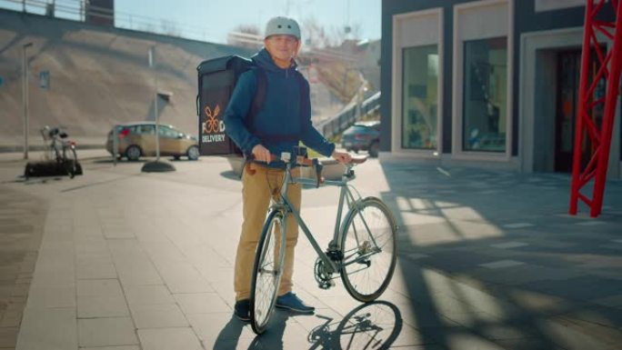 穿着保暖背包和安全帽的英俊快乐送餐男子站在时尚的现代市区的自行车旁边。微笑的快递员递送餐厅订单的肖像